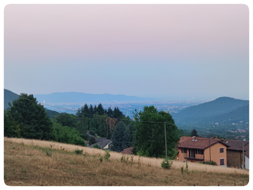 In primo piano, un pascolo in leggera pendenza. L'erba è dorata, bruciata dal sole. Di fronte si vedono alcuni alberi e una casa vicina. Al di là, sullo sfondo, si apre la pianura torinese. Della città sono visibili solo tracce, nascoste dalla nebbia formata dall'afa che aleggia. In lontananza si intravede la collina torinese.