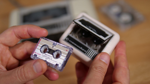 L'immagine mostra una mano che tiene un nastro magnetico Sony MC-650M, con la sua confezione trasparente. Il nastro è piccolo e rettangolare, con la scritta "SONY" e "MC-650M" visibili sulla superficie. La mano tiene il nastro con la punta del dito indice e il pollice, mostrando la sua piccola dimensione. Sullo sfondo, si vede un'apertura di un lettore di nastri, probabilmente un modello Commodore, con la sua parte interna visibile. Sul piano di lavoro, si intravede un altro nastro magnetico in una confezione simile. L'immagine evoca un senso di nostalgia per i media analogici.