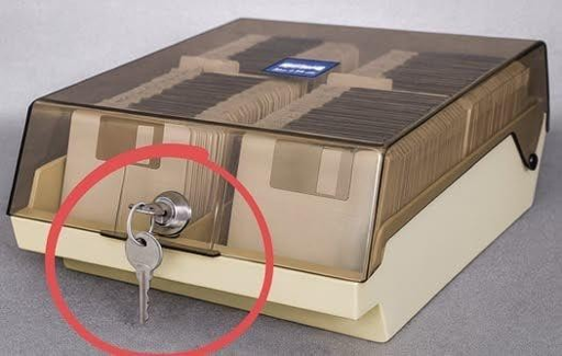 L'immagine mostra un contenitore di floppy ddisk. Il dispositivo è di colore beige con una parte superiore trasparente che permette di vedere i dischi all'interno. La parte superiore è protetta da una copertura di plastica traslucida, che protegge i dischi da polvere e danni. Sul lato anteriore, c'è una serratura con una chiave inserita, evidenziata da un cerchio rosso. La chiave è di metallo e sembra essere la chiave di accesso al contenuto dell'archivio. Sul lato superiore, c'è un piccolo etichettatore blu con la scritta "Kodak", indicando la marca del prodotto. L'archivio è posizionato su una superficie grigia, probabilmente un tavolo o un mobile.

