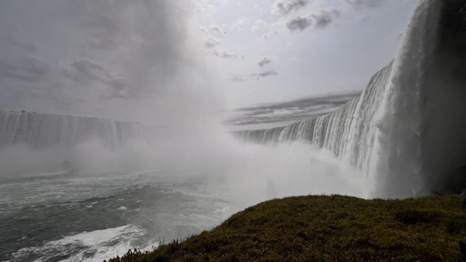 A dramatic view of a large waterfall, with cascading water and mist creating a cloudy atmosphere. The foreground features green grass, while the surrounding cliffs rise sharply, enhancing the scale of the scene.