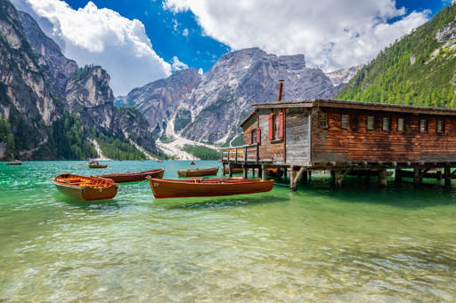 Un’affascinante vista di un lago turchese circondato da montagne scoscese e rocciose con tratti di vegetazione. Nel primo piano, diverse barche in vimini galleggiano in acqua limpida e poco profonda. A destra, una grande casa in legno appoggiata a sillage si estende sull’acqua, con diverse finestre e un piccolo camino. Sullo sfondo si vedono vette montuose drammatiche con macchie di neve sotto un cielo blu brillante con nuvole sparsi bianche. L'acqua trasforma gradualmente dal turchese profondo vicino alle barche a verde più chiaro nelle zone poco profonde.