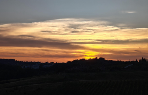 Un tramonto suggestivo dipinge il cielo sopra la silhouette dei colli senesi.
Il Cielo e le Sfumature
Colori caldi: Sfumature intense di giallo oro e arancione acceso si concentrano all'orizzonte.
Sfumature fredde: Le tonalità calde sfumano verso l'alto in tonalità grigio-azzurre e nuvole color crema.
Luce diffusa: I raggi del sole, nascosti dalle colline, illuminano dal basso una vasta coltre nuvolosa.
Il Paesaggio e la Terra
Profili scuri: Le colline toscane creano una linea d'ombra netta e sinuosa contro la luce del cielo.
Elementi tipici: Si intravedono profili di alberi, forse i classici cipressi, e la sagoma di un borgo in lontananza.
Primo piano: I campi e i vigneti in ombra digradano dolcemente, avvolti dal crepuscolo.
