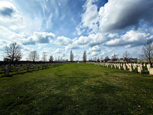 L’immagine mostra un cimitero militare con due file simmetriche di lapidi chiare disposte su un ampio prato verde. Al centro, in lontananza, si erge una grande croce bianca. Il cielo è ampio e dominato da nuvole bianche e grigie sparse, con la luce che filtra creando un’atmosfera serena e silenziosa. Ai lati, dietro le lapidi, ci sono alberi spogli o con poche foglie, tipici di una stagione fredda o di inizio primavera.