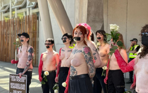 Un gruppo di donne transgender, è in piedi in un'area aperta, probabilmente durante una protesta. Le donne sono nude fino alla vita, con le braccia e le gambe coperte da pantaloni neri. Alcune hanno il viso coperto da nastro di gomma nero, simbolo di silenzio. Ognuna tiene in mano una rosa bianca, simbolo di pace o protesta. Alcune hanno tatuaggi visibili sulle braccia e sul petto. Una donna al centro ha i capelli rossi e un nastro rosa in testa. Sullo sfondo, si vede un cartello con la scritta "THEY IGNORED OUR VOICES REFUSED OUR SUBMISSIONS DID NOT," indicando un messaggio di protesta. La scena è illuminata da una luce solare, suggerendo che l'evento si svolge durante il giorno.