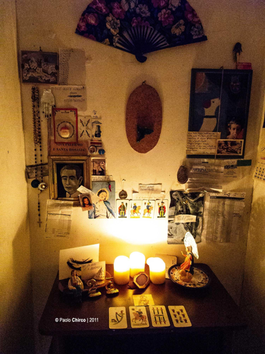 Un altare laico e profano allestito in un angolo di Casa AUT a Cinisi. Su un mobile di legno scuro brillano tre candele accese che illuminano carte da gioco siciliane, piccoli busti e ritratti. La parete sovrastante è densa di icone: una foto d'epoca di un uomo, immagini sacre, un ventaglio aperto a fiori, rosari appesi e vari ritagli di carta. L'atmosfera è calda e carica di simbolismo, trasformando l'ambiente domestico in uno spazio di memoria collettiva.