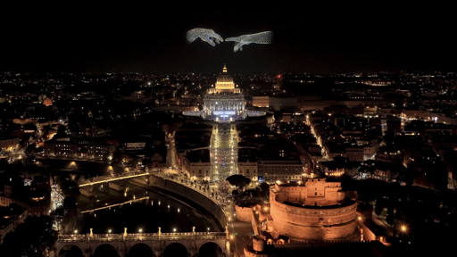 Vista notturna aerea di Roma con un edificio cupolato illuminato al centro, due forme simili a mani illuminate sopra che rappresentano la famosa Creazione di Adamo, e un edificio circolare nell'angolo inferiore destro