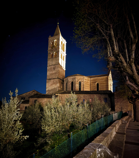 Campanile e chiesa illuminati di notte con cielo stellato, alberi e strada asfaltata.