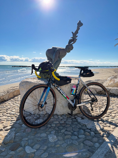 Bicicletta da corsa appoggiata a una statua sulla spiaggia, con il mare e il cielo azzurro sullo sfondo.
