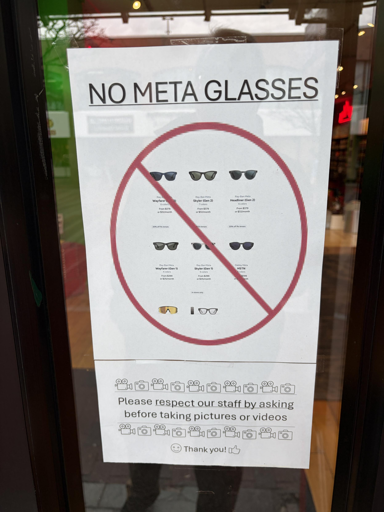 Un cartello bianco affisso su una superficie di vetro. In alto, la scritta in grassetto e maiuscolo recita "NO META GLASSES". Al centro, un grande simbolo di divieto rosso, composto da un cerchio e una barra diagonale, è sovrapposto a diverse immagini di modelli di occhiali Ray-Ban Meta con i relativi prezzi. Nella parte inferiore, una riga di icone di fotocamere e videocamere precede il testo "Please respect our staff by asking before taking pictures or videos". In fondo, un'altra serie di icone è seguita dal testo "😊 Thank you! 👍".