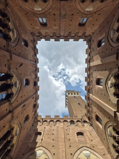 Vista dal basso del cortile interno del Palazzo Pubblico di Siena, con la Torre del Mangia che si staglia contro il cielo nuvoloso.