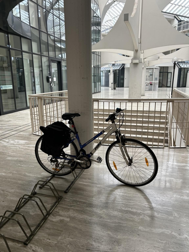 A bicycle is parked on a bike rack inside a modern, spacious building with large glass walls. The interior features marble flooring and a distinctive ceiling design.