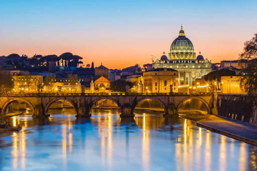 Un panorama di Roma. Un ponte con archi in pietra attraversa il fiume, con luci che illuminano la struttura. Sulla destra, la grande cupola svetta sopra edifici e alberi sotto un cielo che sfuma dal blu all'arancione. Riflessi luminosi di luci gialle sono visibili sulla superficie dell'acqua. 