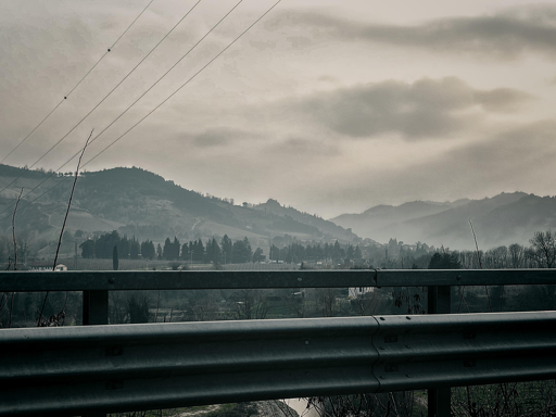 L’immagine mostra un paesaggio collinare in una giornata grigia e nebbiosa. In primo piano c’è un guardrail metallico lungo una strada sopraelevata, mentre sullo sfondo si vedono colline e montagne avvolte nella foschia. Alcuni alberi spogli e conifere punteggiano la vallata, e fili dell’alta tensione attraversano il cielo coperto da nuvole dense e scure. L’atmosfera è malinconica e invernale.