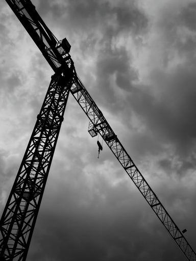 L’immagine mostra una gru da cantiere in silhouette contro un cielo nuvoloso e scuro, creando un’atmosfera drammatica. La struttura metallica della gru si staglia in diagonale, mentre le nuvole dense e grigie sullo sfondo accentuano il contrasto e la sensazione di tensione visiva.