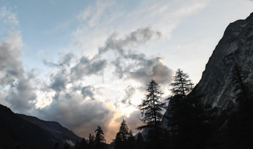 L'immagine mostra un tramonto suggestivo in un paesaggio montano.Il Cielo e la LuceColori caldi: Un chiarore dorato e arancione si diffonde dal basso, dietro le montagne.Sfumature fredde: La parte superiore del cielo sfuma in un azzurro chiaro e limpido.Nuvole dinamiche: Masse nuvolose grigie e bianche circondano il punto in cui il sole tramonta.Contrasti intensi: La luce solare illumina i bordi delle nuvole, creando un forte contrasto con le zone d'ombra.Gli Elementi NaturaliSagome scure: In primo piano spiccano i profili neri di alti alberi, probabilmente larici o abeti.Montagne imponenti: Sul lato destro si erge una parete rocciosa ripida e scura che domina la scena.Orizzonte montano: Sullo sfondo a sinistra si intravedono i profili di altre catene montuose più basse.