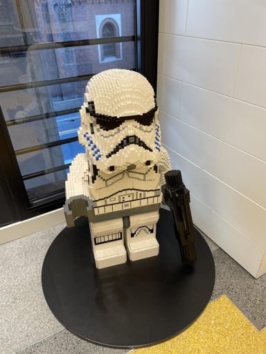 L'immagine mostra una figura di un Stormtrooper, realizzata con mattoncini LEGO, posizionata su un piatto nero. La figura è dettagliata, con una tuta bianca e blu, un elmetto con visiera nera e una pistola LEGO nera in mano. La base su cui è posizionata è circolare e nera, e la figura è collocata in un ambiente interno con un pavimento grigio e una parete bianca. In lontananza, attraverso una finestra, si intravede un edificio in mattoni rossi. La figura è ben illuminata, mettendo in risalto i dettagli e le texture dei mattoncini LEGO.
