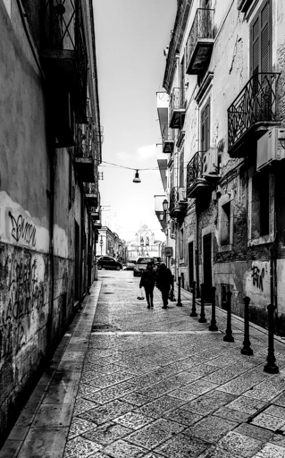 Foto verticale in bianco e nero di una strada stretta del centro storico di Foggia. Il pavimento è in pietra, visto in prospettiva verso il fondo della via. Ai lati ci sono palazzi alti con balconi e condizionatori, alcuni muri sono segnati da graffiti. Al centro dell’inquadratura due persone camminano una accanto all’altra, una tiene in mano una busta. In fondo alla strada si vedono alcune auto parcheggiate e la facciata di un edificio più luminoso contro il cielo chiaro.