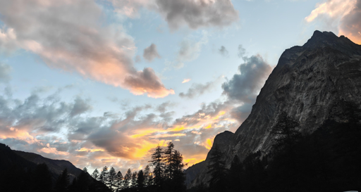 L'immagine cattura un suggestivo tramonto in Val Veny, dominato dal contrasto tra la maestosità della roccia e i colori caldi del cielo.Ecco i dettagli principali della scena:Il cielo e le nuvole: La parte superiore è attraversata da nuvole sparse, illuminate dagli ultimi raggi di sole. Sfumature che vanno dal rosa tenue all'arancione dorato si mescolano con il grigio e l'azzurro del cielo, creando un forte dinamismo visivo.Le montagne: Sulla destra si staglia l'imponente e ripida parete rocciosa di una montagna, lasciata in gran parte in ombra, che accentua il senso di verticalità tipico del paesaggio alpino.Il profilo della valle: In basso, le silhouette scure degli alberi (principalmente conifere) e i profili delle creste montuose circostanti incorniciano la luce del sole, che scompare dietro le vette lasciando un bagliore giallo e arancione intenso.