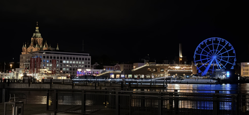 Night view of Helsinki’s waterfront from Kauppatori on 5 September 2026 at 21:54. Across the dark harbour, the Allas Sea Pool complex is illuminated with warm lights, beside a large Ferris wheel glowing electric blue. The ornate tower of Uspenski Cathedral rises above the buildings on the left, while rows of lights along the quayside shimmer in reflections on the water beneath a black night sky.