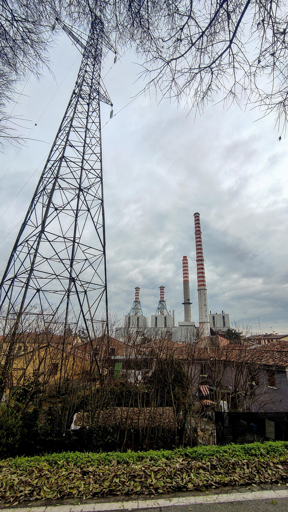 Vista esterna di un’area industriale con un grande traliccio elettrico in primo piano e, sullo sfondo, una centrale con quattro alte ciminiere a strisce rosse e bianche. In basso si vedono case con tetti marroni e alberi spogli, sotto un cielo nuvoloso.