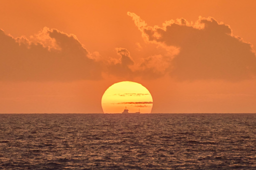 Un tramonto sull’oceano presenta un grande, luminoso sole giallo-arancio all’orizzonte, parzialmente oscurato da leggere nuvole. Il cielo mostra tonalità calde di arancione e rosa, con nuvole sparpagliate che mostrano bordi più leggeri dalla luce solare. Il mare sottostante ha una superficie scura e ruvida con onde delicate. Una piccola nave è visibile all’orizzonte a destra del sole.