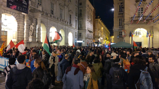 Piazza del centro di Torino con palazzi antichi e porticati illuminati. La piazza è piena di manifestanti con bandiere della Palestina e di partiti e associazioni sindacali.