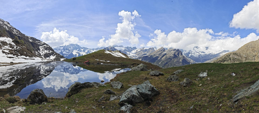 L'immagine mostra un panorama montano ad ampio raggio (grandangolare o panoramica) caratterizzato da un laghetto alpino e vette innevate sullo sfondo.Elementi PrincipaliIl laghetto alpino: Situato sulla sinistra, riflette in modo nitido il cielo azzurro, le nuvole bianche e le montagne circostanti.Il terreno: In primo piano e sulla destra si estende un pendio erboso intervallato da rocce grigie sparse e chiazze di neve in via di scioglimento.Le montagne: Sullo sfondo si staglia una catena montuosa imponente con vette parzialmente innevate che definiscono l'orizzonte.Il cielo: Di colore azzurro intenso, è attraversato da nuvole cumuliformi bianche e soffici