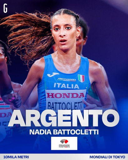 Atleta italiana Nadia Battocletti in gara nei 10.000 metri, con canotta azzurra della nazionale e pettorale con sponsor Honda. Testo in sovrimpressione: ‘Argento Nadia Battocletti’ per la medaglia d’argento ai Mondiali di atletica di Tokyo 2025.

