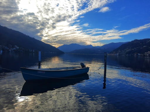Un suggestivo panorama di un lago sereno con un piccolo battello blu appoggiato in acqua calma, legato a due pali in legno. Il battello ha un motore visibile alla sterna e un numero di telefono recante la scritta “F 3059”. L’acqua riflette il cielo, caratterizzato da miriadi di macchie blu luminose e nuvole irregolari con raggi di sole che filtrano attraverso. Sullo sfondo, si ergono montagne e colline spoglie, con vegetazione rada e piccoli edifici. L'ambientazione è tranquilla, con il cielo e il paesaggio che creano uno scenario all'aria aperta pacifico.