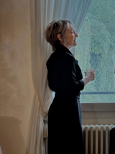 A person stands by a window, holding a cup and smiling. She has short, styled hair and is wearing a black outfit. Light filters through sheer curtains, illuminating the scene. Green foliage is visible outside the window.
