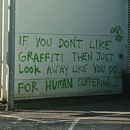 L'immagine mostra un muro bianco su cui è scritto in verde un messaggio di graffiti. Il testo recita: "IF YOU DON'T LIKE GRAFFITI THEN JUST LOOK AWAY LIKE YOU DO FOR HUMAN SUFFERING..." (Se non ti piace il graffiti, allora guarda via come fai per il sofferente umano...). Il messaggio è scritto in lettere maiuscole e ha un aspetto informale, tipico del graffiti. Sul lato destro del muro, si nota un piccolo simbolo grafico. Il muro è situato in un'area aperta, con un pavimento asfaltato e una barriera metallica visibile in primo piano.